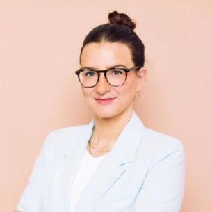 Eine Person mit Brille und weißem Blazer vor hellem Hintergrund